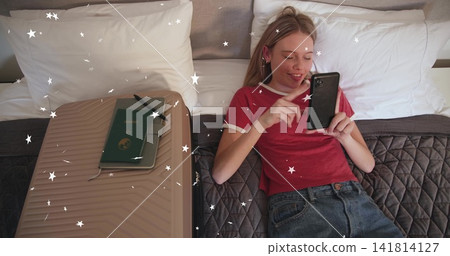 Reclining woman in red tee and jeans holding smartphone, scrolling on bed, with suitcase passport Reclining woman in red tee and jeans holding smartphone, scrolling on bed, with suitcase passport 141814127