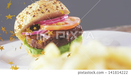 Sesame-seed hamburger sitting on white plate at tabletop, with gold star confetti and blurred fries Sesame-seed hamburger sitting on white plate at tabletop, with gold star confetti and blurred fries 141814222