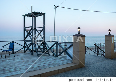 Seaside pier with metal railings, stone pillars and an observation tower overlooking calm blue water, creating a peaceful coastal scene with maritime architecture and open horizon view Seaside pier with metal railings, stone pillars and an observation tower overlooking calm blue water, creating a peaceful coastal scene with maritime architecture and open horizon view 141814918
