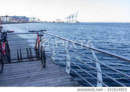 Wooden seaside pier with metal railings extending along blue water, featuring benches and bicycles, with distant harbor cranes creating a peaceful coastal urban landscape view Wooden seaside pier with metal railings extending along blue water, featuring benches and bicycles, with distant harbor cranes creating a peaceful coastal urban landscape view 141814923