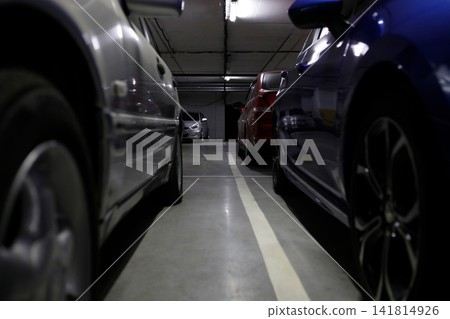 Low angle view between parked cars inside an underground parking garage, showing narrow driving lane, concrete floor, artificial lighting and modern urban transport infrastructure Low angle view between parked cars inside an underground parking garage, showing narrow driving lane, concrete floor, artificial lighting and modern urban transport infrastructure 141814926