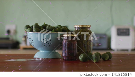 Displaying blue colander filled with green fruits on wooden countertop, with jam and pickle jars Displaying blue colander filled with green fruits on wooden countertop, with jam and pickle jars 141815814