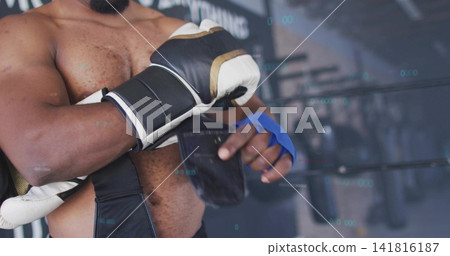 Adjusting shirtless boxer securing black strap at boxing gym near ring, with blue wraps and glove Adjusting shirtless boxer securing black strap at boxing gym near ring, with blue wraps and glove 141816187