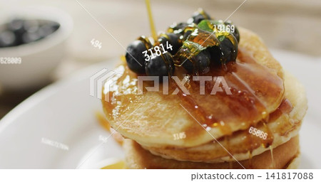 Pouring syrup over three-layer pancake stack on kitchen table, with blueberries and white bowl Pouring syrup over three-layer pancake stack on kitchen table, with blueberries and white bowl 141817088