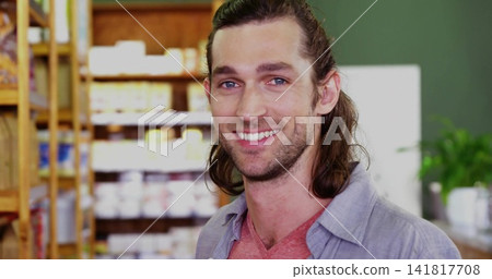 Smiling mid-adult man standing and facing camera in small shop, wearing gray shirt, wooden shelf Smiling mid-adult man standing and facing camera in small shop, wearing gray shirt, wooden shelf 141817708