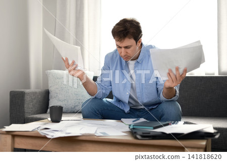 Man under stress holding unpaid utility bills at home. Frustrated by rising household expenses and financial debt, he faces an economic inflation crisis. Man under stress holding unpaid utility bills at home. Frustrated by rising household expenses and financial debt, he faces an economic inflation crisis. 141818620