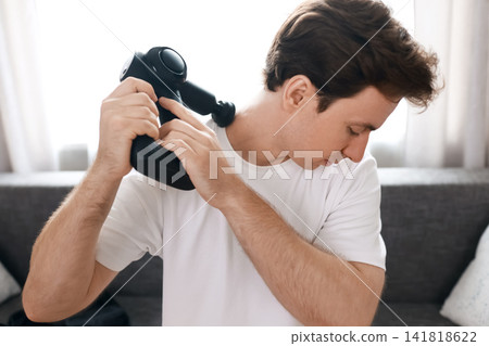 Man using a percussion massage gun on his neck and shoulder for pain relief and muscle tension recovery. Selfcare therapy and myofascial relaxation at home. Man using a percussion massage gun on his neck and shoulder for pain relief and muscle tension recovery. Selfcare therapy and myofascial relaxation at home. 141818622