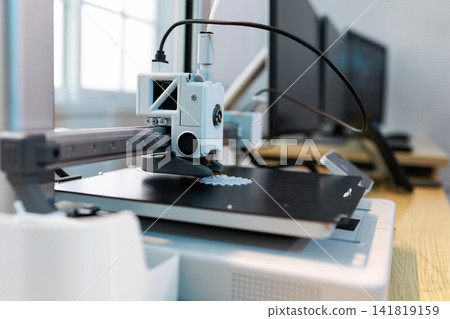 Desktop 3d printer printing a small model on a work table in a modern office, with blurred monitors and tools in the background. Desktop 3d printer printing a small model on a work table in a modern office, with blurred monitors and tools in the background. 141819159
