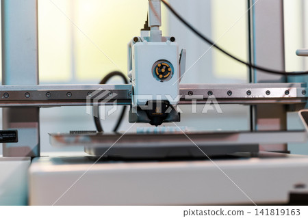 Close up of a 3d printer head working on a build plate in a workshop, with blurred cables and machine parts in the background. Close up of a 3d printer head working on a build plate in a workshop, with blurred cables and machine parts in the background. 141819163