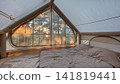 Cozy tent interior with quilted sleeping pad, mesh window, sunrise over pine forest, warm golden light filtering through fabric walls and soft morning shadows across camp bed 141819441