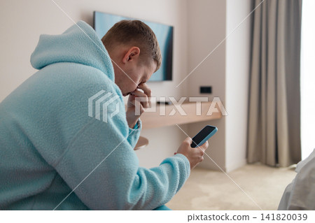 Thoughtful young man using smartphone sitting on bed in room Thoughtful young man using smartphone sitting on bed in room 141820039