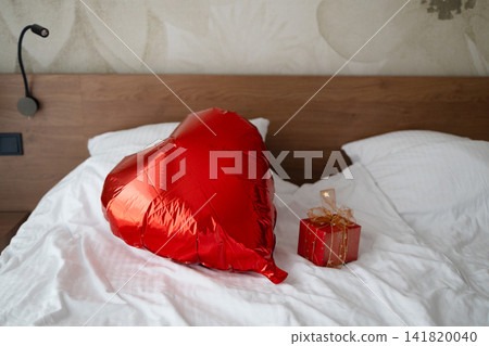 Red heart shaped balloon and gift box on bed Red heart shaped balloon and gift box on bed 141820040