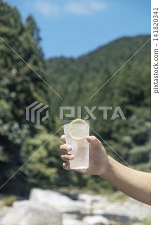 A hand holding a lemon-flavored sparkling water bottle against a backdrop of blue sky and green mountains. A hand holding a lemon-flavored sparkling water bottle against a backdrop of blue sky and green mountains. 141820341