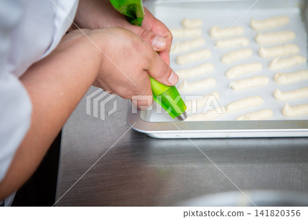 Pastry chef piping neat rows of dough onto metal baking tray in bright stainless steel kitchen, preparing uniform sweets for commercial bakery production with steady precision Pastry chef piping neat rows of dough onto metal baking tray in bright stainless steel kitchen, preparing uniform sweets for commercial bakery production with steady precision 141820356