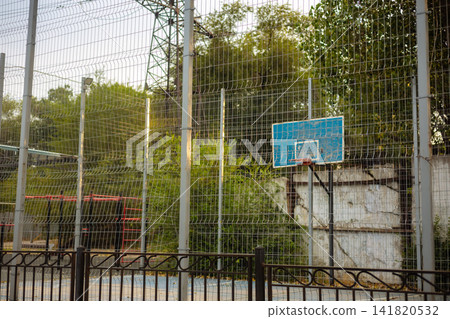Outdoor basketball court behind wire mesh fence with blue backboard and trees Outdoor basketball court behind wire mesh fence with blue backboard and trees 141820532