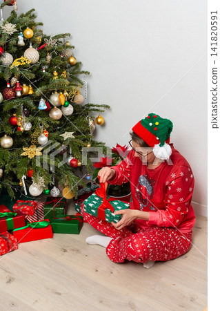 A 52-year-old woman sits on the floor next to a Christmas tree. She is smiling and holding a wrapped gift while wearing a festive hat. It is Christmas Day and there are many presents around A 52-year-old woman sits on the floor next to a Christmas tree. She is smiling and holding a wrapped gift while wearing a festive hat. It is Christmas Day and there are many presents around 141820591