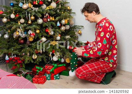 A 53 year old man sits near a decorated Christmas tree on Christmas Day. He is opening a gift while wearing a festive outfit and a holiday hat. Family and joy fill the space around him A 53 year old man sits near a decorated Christmas tree on Christmas Day. He is opening a gift while wearing a festive outfit and a holiday hat. Family and joy fill the space around him 141820592