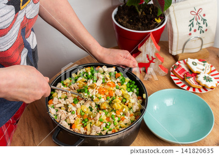 A woman mixes ingredients for Olivier salad, Russian salad, a traditional Ukrainian or Russian Christmas or New Year dish in a kitchen. She sits at a wooden table with a bowl and cutting board nearby A woman mixes ingredients for Olivier salad, Russian salad, a traditional Ukrainian or Russian Christmas or New Year dish in a kitchen. She sits at a wooden table with a bowl and cutting board nearby 141820635