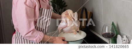 In a cozy kitchen, a 53-year-old man washes chicken in the sink while getting ready for a pre-Christmas dinner. He has potatoes and fruits on the table beside him, banner In a cozy kitchen, a 53-year-old man washes chicken in the sink while getting ready for a pre-Christmas dinner. He has potatoes and fruits on the table beside him, banner 141820665
