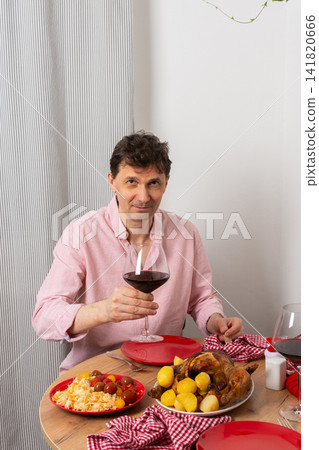 A man is cooking dinner for Christmas by baking chicken with potatoes in his kitchen. He is sitting at the table, ready to enjoy the meal with a glass of wine A man is cooking dinner for Christmas by baking chicken with potatoes in his kitchen. He is sitting at the table, ready to enjoy the meal with a glass of wine 141820666