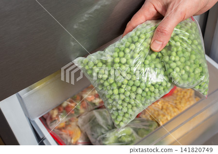 A man opens the refrigerator to take out a bag of frozen green peas. He is in the kitchen, preparing food for a meal in the afternoon. The refrigerator is organized with several bags inside 141820749