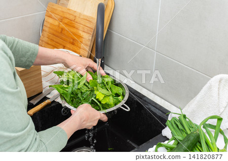 Hands are holding a colander filled with fresh organic local vegetables under running water in a kitchen sink. This is done to clean the produce before cooking Hands are holding a colander filled with fresh organic local vegetables under running water in a kitchen sink. This is done to clean the produce before cooking 141820877