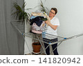 A woman is taking laundry out of the dryer and putting it into a basket. She is in a simple room with plants nearby. Her focus is on the task at hand 141820942