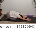 A 52-year-old woman practices yoga in her living room. She lies on a mat with her arms stretched forward. The setting shows a home environment with plants nearby 141820943
