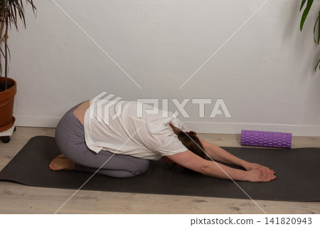 A 52-year-old woman practices yoga in her living room. She lies on a mat with her arms stretched forward. The setting shows a home environment with plants nearby A 52-year-old woman practices yoga in her living room. She lies on a mat with her arms stretched forward. The setting shows a home environment with plants nearby 141820943