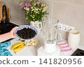 A woman stands in a kitchen holding a bowl of currants above a blender. Bowls of sliced bananas are nearby. Flowers are displayed on the counter, adding to the cooking scene 141820956