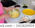 In a kitchen, a woman mixes ingredients to make sour cream sauce for a shortbread pie. Fresh strawberries, currants, and blueberries are ready for the pie filling 141820959