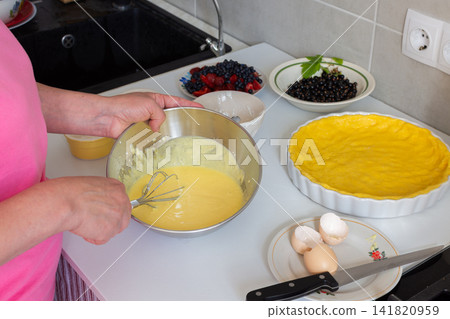 In a kitchen, a woman mixes ingredients to make sour cream sauce for a shortbread pie. Fresh strawberries, currants, and blueberries are ready for the pie filling In a kitchen, a woman mixes ingredients to make sour cream sauce for a shortbread pie. Fresh strawberries, currants, and blueberries are ready for the pie filling 141820959
