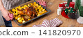 A man preparing for a pre-Christmas dinner places a tray of baked chicken and potatoes on a wooden table. A glass of red wine sits nearby as he focuses on the meal, banner 141820970