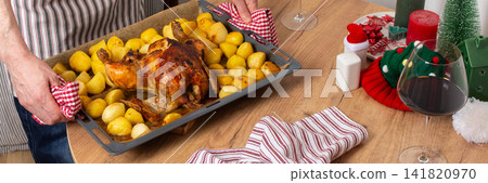 A man preparing for a pre-Christmas dinner places a tray of baked chicken and potatoes on a wooden table. A glass of red wine sits nearby as he focuses on the meal, banner A man preparing for a pre-Christmas dinner places a tray of baked chicken and potatoes on a wooden table. A glass of red wine sits nearby as he focuses on the meal, banner 141820970
