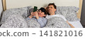 A married couple wakes up in bed on a typical morning. The woman is using a phone while the man lies next to her. They share a cozy moment in their bedroom, banner 141821016
