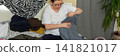 A 52-year-old woman is sorting clean laundry on a bed. She holds a pair of pants in one hand while looking at the clothes around her. A plant is seen in the corner of the room, banner 141821017