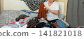 A woman, age 52, sits on her bed in a bedroom while sorting through clean laundry. She holds a piece of clothing in her hands as she organizes her belongings on the bed, banner 141821018
