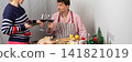 A man is baking chicken with potatoes for a pre-Christmas dinner. He is talking to a woman while cooking. They are in a cozy kitchen with festive decorations and wine, banner 141821019