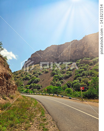 Scenic mountain road winds through sunlit hills with green vegetation Scenic mountain road winds through sunlit hills with green vegetation 141821554