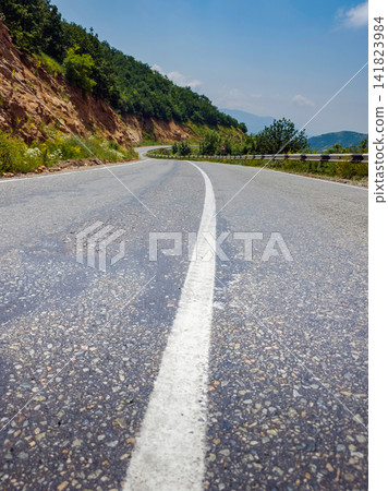 Winding rural road with white line, surrounded by trees and hills under blue sky Winding rural road with white line, surrounded by trees and hills under blue sky 141823984