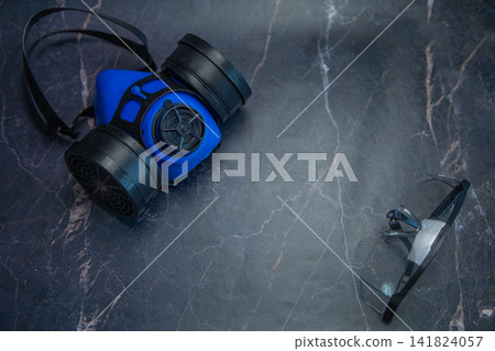 A blue respirator and goggles are spaced apart on a dark marble table. Concept, minimalism in labor protection, respiratory and visual protection, free space for text, safety. High quality photo A blue respirator and goggles are spaced apart on a dark marble table. Concept, minimalism in labor protection, respiratory and visual protection, free space for text, safety. High quality photo 141824057