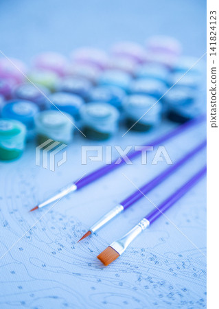 Three brushes with purple handles lie on a numbered canvas, and blurry paint jars are visible in the background. Concept of preparation for creativity, artistic tools, focus on the process. High Three brushes with purple handles lie on a numbered canvas, and blurry paint jars are visible in the background. Concept of preparation for creativity, artistic tools, focus on the process. High 141824123