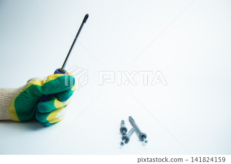 A hand with a construction glove holds a screwdriver on a white background, next to it are self-tapping screws with space for text. Concept of repair, handyman, tools, construction, installation. High A hand with a construction glove holds a screwdriver on a white background, next to it are self-tapping screws with space for text. Concept of repair, handyman, tools, construction, installation. High 141824159