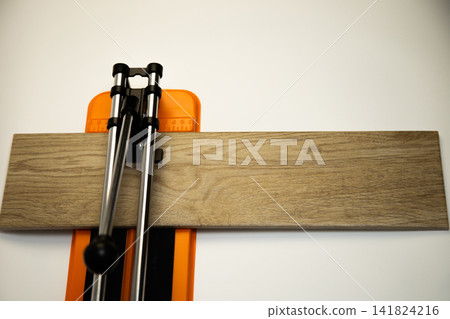 A top view of an orange mechanical tile cutter with a metal river, on which ceramic tiles of wooden colors lie sideways on a white background. The concept of renovation, tiling, construction work A top view of an orange mechanical tile cutter with a metal river, on which ceramic tiles of wooden colors lie sideways on a white background. The concept of renovation, tiling, construction work 141824216