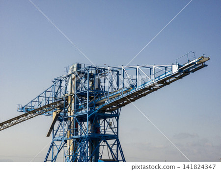 Massive blue steel industrial conveyor crane structure stands against clear sky for bulk material handling Massive blue steel industrial conveyor crane structure stands against clear sky for bulk material handling 141824347