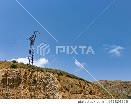 Power line tower rises above arid hillside against bright blue sky Power line tower rises above arid hillside against bright blue sky 141824526