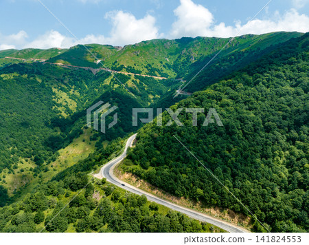 Winding asphalt road snakes through forested mountains under bright sky Winding asphalt road snakes through forested mountains under bright sky 141824553