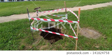 Deep dark hole in the ground cordoned with red and white caution tape on wooden stakes. Deep dark hole in the ground cordoned with red and white caution tape on wooden stakes. 141824758