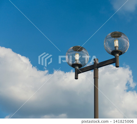A retro double-headed streetlamp stands against a backdrop of blue sky and white clouds, an outdoor lighting fixture and a skyscape. A retro double-headed streetlamp stands against a backdrop of blue sky and white clouds, an outdoor lighting fixture and a skyscape. 141825006