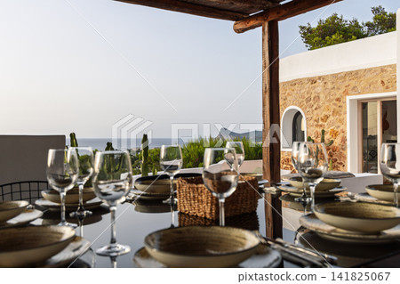 Outdoor dinner table on Ibiza, wide villa dining scene with stone facade and sea view. Outdoor dinner table on Ibiza, wide villa dining scene with stone facade and sea view. 141825067